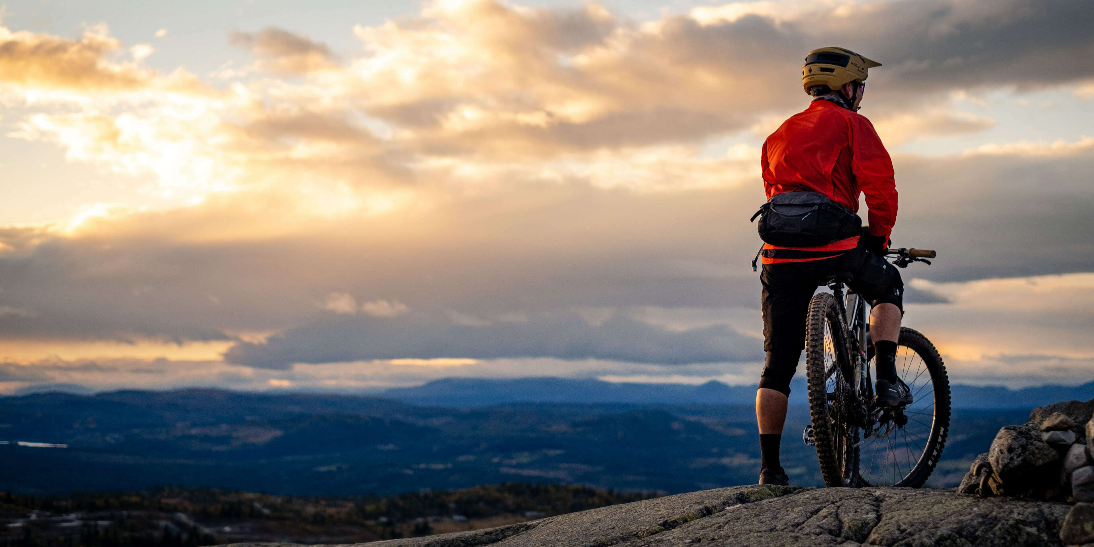 Mann på sykkel på fjellet som ser utover fjellandskapet i solnedgang. Foto.